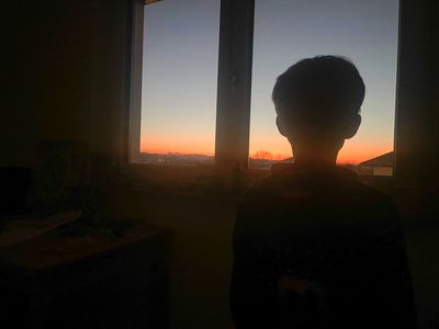 A photo of Emil from behind while he is watching the evening sky through the window of his room.